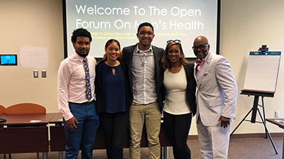 Five people stand in front of presentation that says &quot;Welcome to the open forum on men's health&quot;